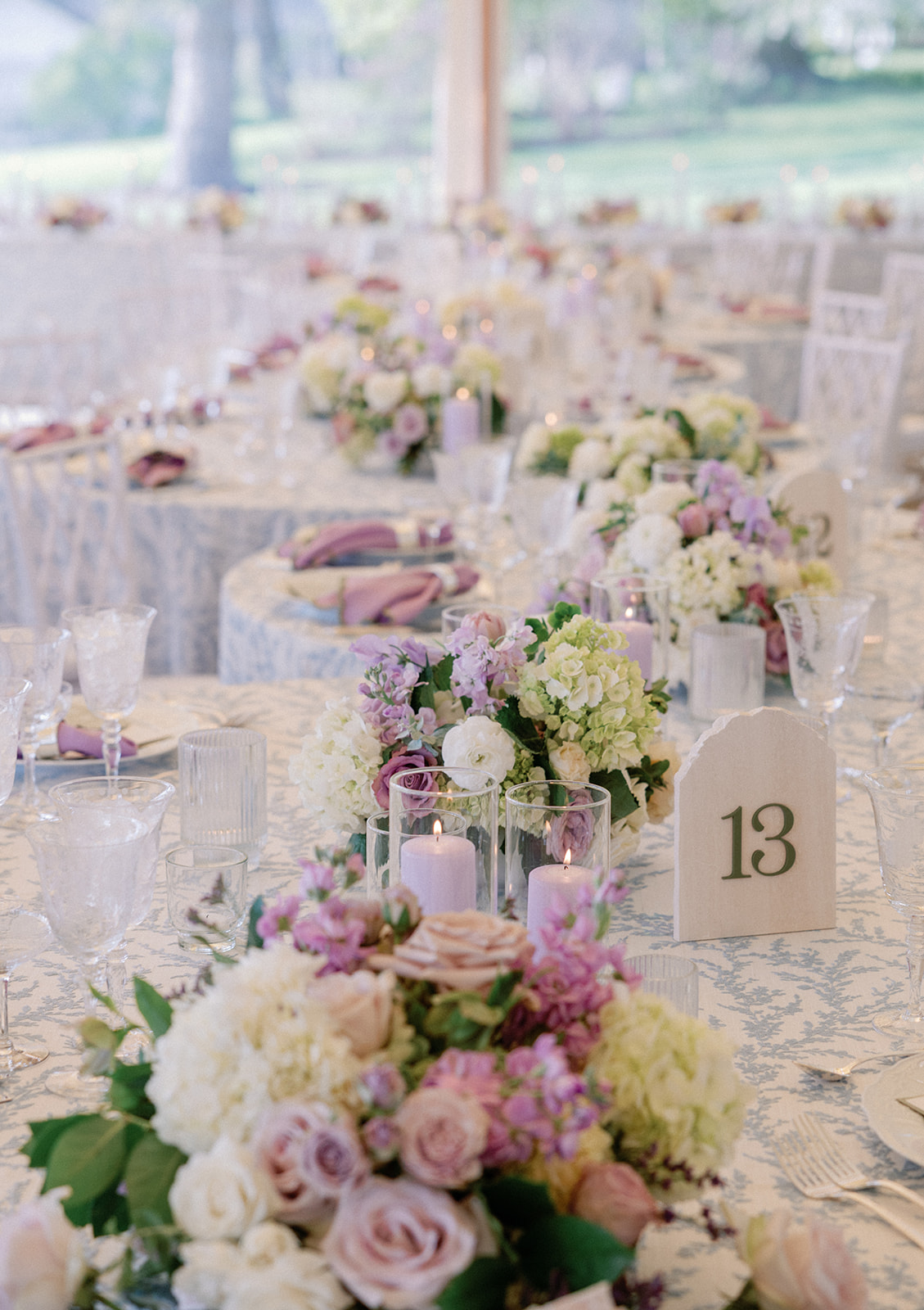 Close-up of a table setting with a white, pink, and lavender floral centerpiece beside a lit taper candle in a glass cylinder, next to a wooden table number sign reading '13.'