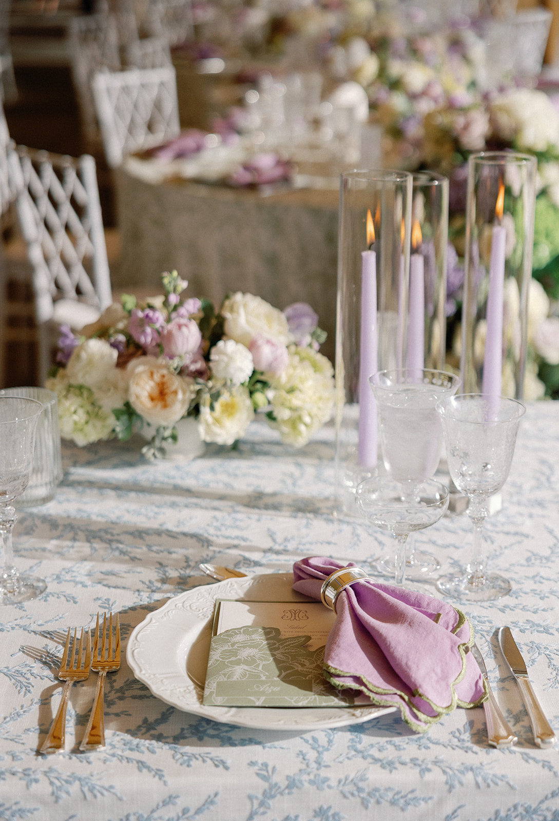 Close-up place setting with a lavender napkin in a silver ring resting on a green floral menu card, gold flatware, and crystal glassware with a lit lavender taper candle, a floral centerpiece blurred behind.