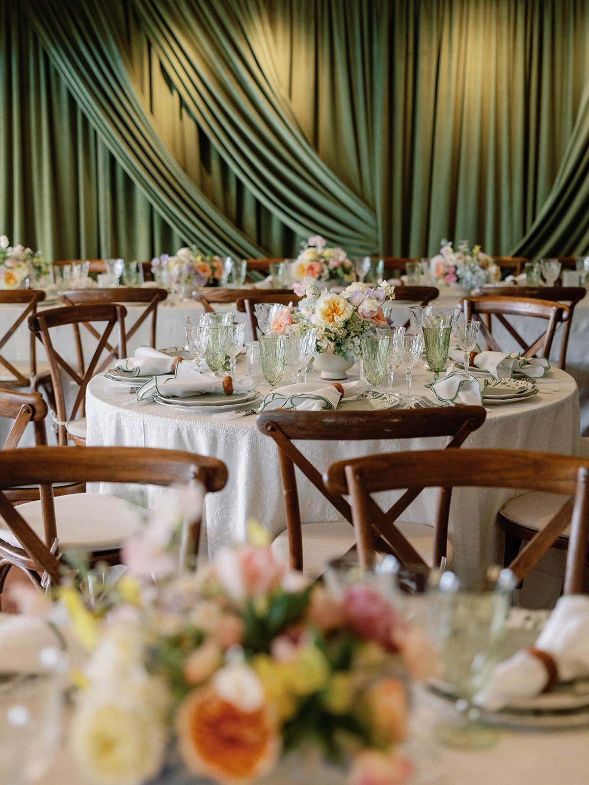 Close-up of a round table setting with green fringe-edged napkins in wooden rings, crystal glassware, and a pastel floral centerpiece, framed by blurred flowers in the foreground.